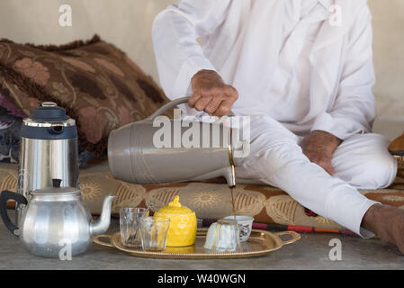 Bédouins arabes homme est assis sur le sol et se verse du thé ou du café à partir de la théière dans la tasse blanche en tente bédouine.arabe traditionnel blanc des vêtements. Banque D'Images