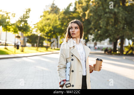 Portrait of cute woman wearing coat grave de boire du café à emporter et holding clipboard en marchant on city street Banque D'Images