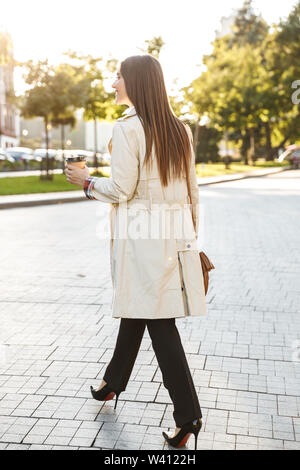 Photo de l'arrière de brunette woman wearing coat de boire du café à emporter tout en marchant on city street Banque D'Images