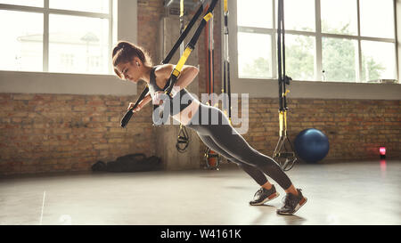 Travailler dur. Vue latérale du jeune femme athlétique avec corps parfait dans les vêtements de sport faisant pousser ups avec trx sangles remise en forme dans la salle de sport. Le sport professionnel. Formation TRX. Workout Banque D'Images