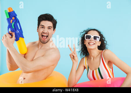 Cheerful young couple isolés sur fond bleu, portant l'anneau gonflable, tenant les pistolets à eau Banque D'Images