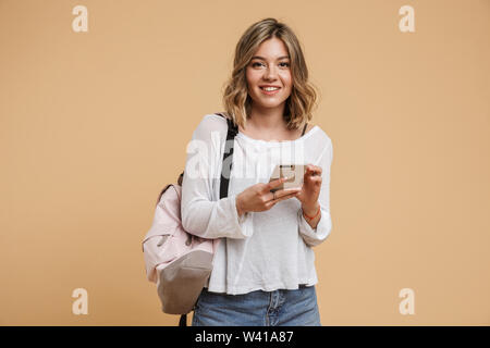 Image de l'adolescente blonde wearing backpack using cellphone and smiling at camera isolé sur fond beige Banque D'Images