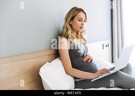 Young pregnant blonde woman relaxing on bed at home, à l'aide d'un ordinateur portable Banque D'Images