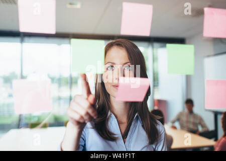 Portrait d'une femme à la recherche professionnelle à un Post-it collé sur un mur de verre dans le bureau Banque D'Images