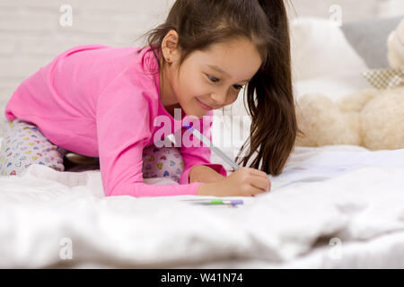 Funny little girl drawing pictures while lying on bed. Peinture pour enfants à la maison Banque D'Images