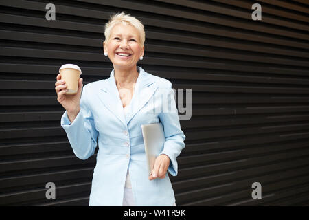 Mature businesswoman with cheveux courts portant costume bleu holding laptop dans sa main et de boire du café à partir de gobelet jetable en plein air Banque D'Images