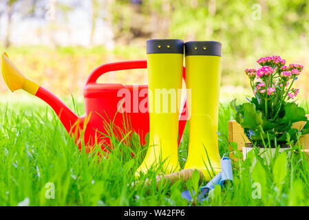 Bottes en caoutchouc jaune, rouge arrosoir et outils pour planter des fleurs sur une pelouse verte close-up Banque D'Images