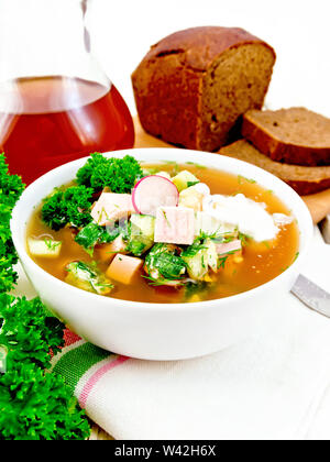Soupe froide d'okroshka, pomme de terre, saucisses, oeufs, concombres, radis verts et kvass dans un bol blanc sur une serviette, pain et verre à boire sur planche de bois ba Banque D'Images