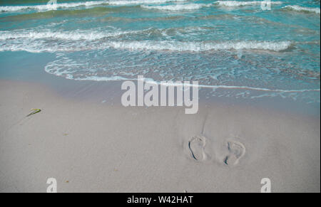 Des traces de pas dans le sable sur la plage, partiellement lavée par les vagues. Maison de vacances sur la plage de sable blanc et mer bleue. Banque D'Images