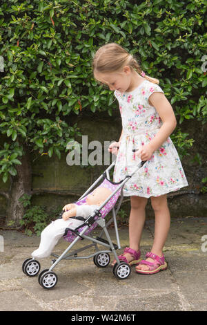 Enfant de trois ans, jeune fille en jolie robe, cheveux blonds en queue de cochon jouant à la poupée dans la poussette jouet, à l'extérieur dans le jardin, UK Banque D'Images