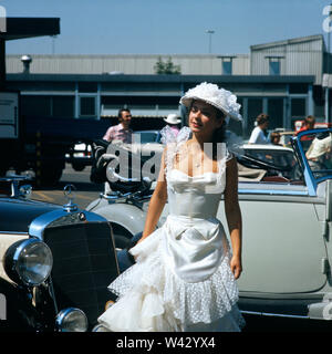 Eine junge Frau in einem weißer bär Kleid steht die zwei Oldtimern auf einer auto de München à den 1980er Jahren. Une jeune femme en robe blanche se tient entre deux voitures sur une voiture à Munich dans les années 80. Banque D'Images