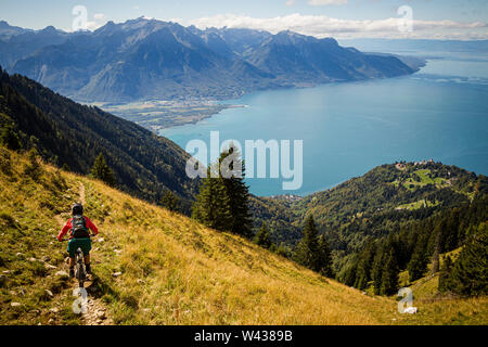 Une femme monte un VTT le long d'un étroit sentier à travers un alpage herbeux raide au-dessus de Montreux et le Lac Léman. Banque D'Images