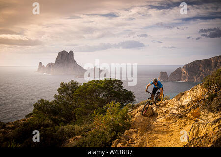 Un homme monte un VTT le long d'un sentier rocheux sur la côte ouest d'Ibiza. Es Vedrá Island et la Méditerranée sont à l'arrière-plan. Banque D'Images