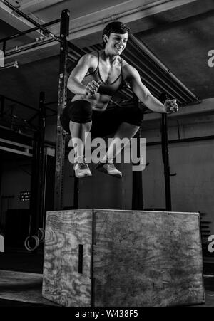 Une femme aux cheveux courts avec fait abs fort fort les sauts dans une salle de sport. L'athlète féminine est de sourire et de sauter sur la boîte. Noir et blanc. Banque D'Images