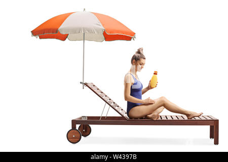 Tourné sur toute la longueur d'une jeune femme l'application d'une crème solaire et stting sur un transat avec parasol isolé sur fond blanc Banque D'Images