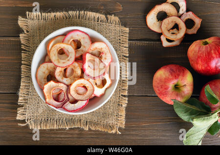 Anneaux de pommes séchées dans un bol blanc sur une table de bois et des pommes fraîches, vue du dessus Banque D'Images