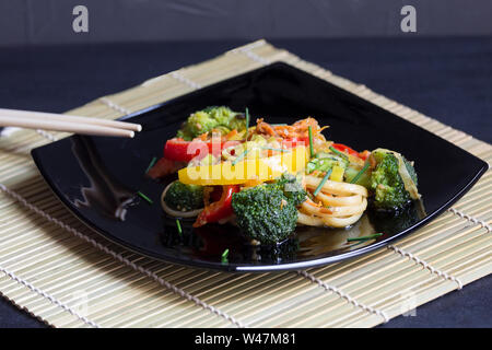 La nourriture asiatique, nouilles Udon noire sur une assiette de légumes et sauce de soja sur un tapis de bambou, fond noir Banque D'Images