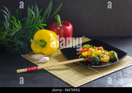 La nourriture asiatique, nouilles sautés avec légumes, sauce soja, poivrons et les ingrédients sur un tapis de bambou, fond noir Banque D'Images