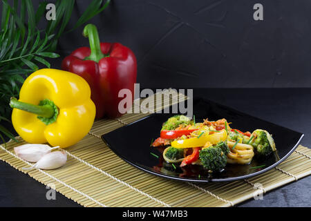 Les nouilles Udon aux légumes sur une plaque noire avec de la sauce soja et d'ingrédients sur un tapis de bambou, fond noir Banque D'Images