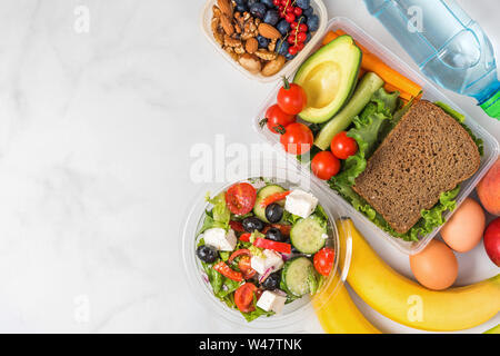 Boîtes à lunch santé avec sandwich, salade grecque, des légumes, des noix, des baies, des bananes, des œufs, de l'avocat, des pêches et de l'eau sur fond blanc. Vue de dessus avec Banque D'Images