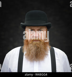 Portrait d'un homme barbu stern entre accolades et bowler hat fixant intensément à l'appareil photo Banque D'Images