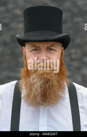 Homme barbu d'âge moyen dans la région de top hat et accolades regardant la caméra avec un sourire amical sur un fond gris Banque D'Images