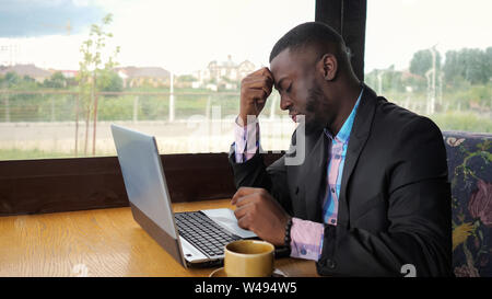 Afro american businessman essaie de les taper sur ordinateur assis dans le café. Homme noir travaille sur l'ordinateur portable, mais il est cassé et il est nerveux et frapper sur les touches. Il a un des problèmes d'ordinateur. La batterie est faible. Banque D'Images