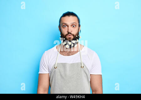 Surpris l'homme, jardinier fleuriste,avec beaucoup de fleurs sur sa barbe en tablier gris en regardant la caméra. isolé sur fond bleu. Banque D'Images