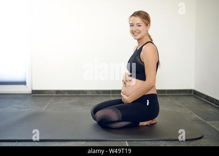 Happy pregnant woman doing exercises à genoux sur un tapis de yoga qui serre son ventre à deux mains tout en souriant à l'appareil photo Banque D'Images