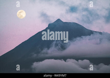 Le mont Pico volcano versant ouest vue de l'océan durant la pleine lune avec sommet en nuages après le coucher du soleil avec ciel rose, du point de vue de l'île de Faial en Açores Banque D'Images