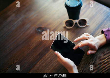 Sommaire des mains des femmes, défilement de l'écran du téléphone mobile. Une tasse de café et verres à côté. Banque D'Images