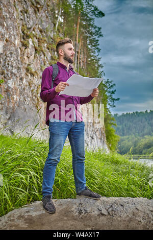 L'éco-tourisme, un jeune homme barbu avec un sac à dos sur son dos, debout sur un rocher avec une carte topographique dans ses mains, et d'explorer le voisinage, Banque D'Images