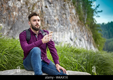 Un jeune homme barbu de blancs ethnos est titulaire d'un smartphone dans ses mains, au cours d'aventures sur un sentier de randonnée, il se repose assis sur une pierre dans l'ecosy Banque D'Images