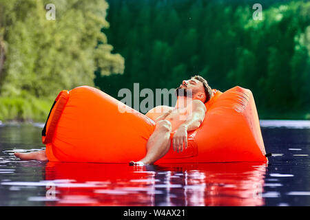 Un jeune homme est allongé dans une chaise longue orange gonflable flottant à la surface de l'eau d'une rivière de la forêt. Camping au cours de l'éco-tourisme. Banque D'Images