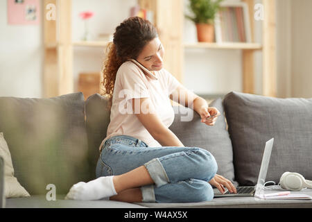 Content jeune femme moderne en tenue décontractée assis sur un canapé et à l'aide d'ordinateur portable tout en commande en ligne et l'appel de l'opérateur de site web Banque D'Images