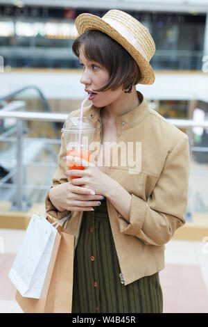 Pensive woman in straw hat holding de sacs en papier et de boire du jus à partir de la paille dans mall Banque D'Images