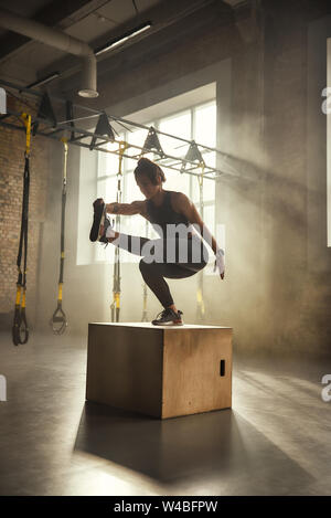 Devenir le meilleur. Vue latérale du sportswear femme athlétique en faisant s'accroupir et de formation les jambes en se tenant sur le boîte en bois à la salle de sport. Le sport professionnel. Formation TRX. Workout Banque D'Images