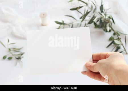 Mariage féminin, maquette de carte de vœux d'anniversaire scène avec woman's hand holding blank carte papier. Arrière-plan flou avec des branches d'arbres d'olive et de soie Banque D'Images