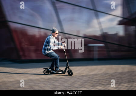 Jeune homme dans des vêtements décontractés sur electric scooter de coup on city street in motion blur contre l'immeuble de bureaux moderne en verre Banque D'Images