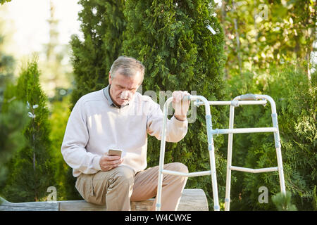 Portrait of senior man with walker reposant sur banc de parc en utilisant smartphone, copy space Banque D'Images