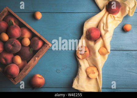 Pêches et abricots mûrs récoltés dans une boîte en bois, vue ci-dessus sur un tableau bleu et jaune une serviette de cuisine. Mise à plat de fruits frais dans un fort. Banque D'Images