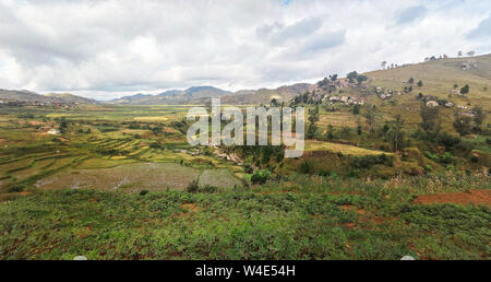 Paysage typique de Madagascar dans la région près de Ambositra petites collines couvertes d'herbe verte et les buissons, maisons d'argile rouge et humide près de champs de riz. Banque D'Images