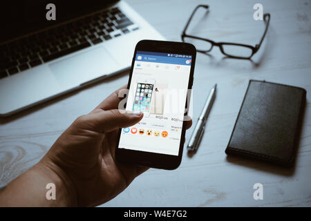 Nizhyn, Ukraine/ juillet-16-2019 : Young man looking at facebook app page sur smartphone moderne. Ordinateur portable, lunettes, stylo et portable sur l'arrière-plan. Me Social Banque D'Images