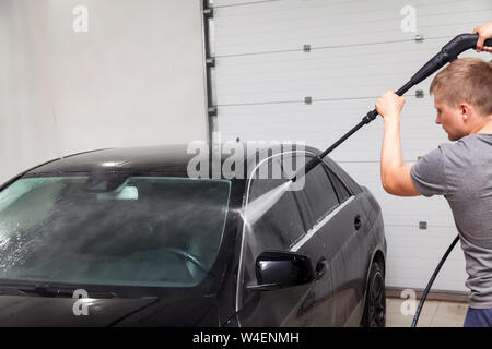 Un travailleur masculin se lave une voiture noire avec un appareil à haute pression où l'eau et élimine la mousse et les produits chimiques dans un delinging véhicule Travaux publics Banque D'Images