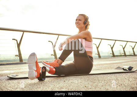 Heureux matin. Athlète handicapée positive woman avec prothèse en faisant les exercices de yoga et d'écouteurs en souriant, assis sur le pont. Mobilité sport concept. La motivation. Mode de vie sain Banque D'Images