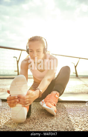 L'échauffement. Photo verticale positive et de belle femme dans la musique et écoute au casque s'étendant sa jambe avec une prothèse en position assise sur le pont. Mobilité sport concept. La motivation. Mode de vie sain Banque D'Images