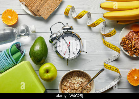 Vue du dessus de l'alimentation nourriture près de ruban à mesurer, réveil et d'équipement de sport sur fond blanc en bois Banque D'Images