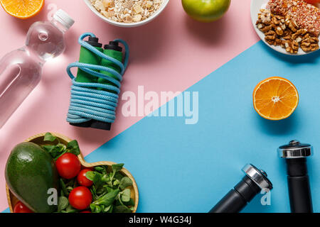 Vue supérieure de l'alimentation l'alimentation, l'eau, la corde à sauter et les haltères sur fond bleu et rose Banque D'Images