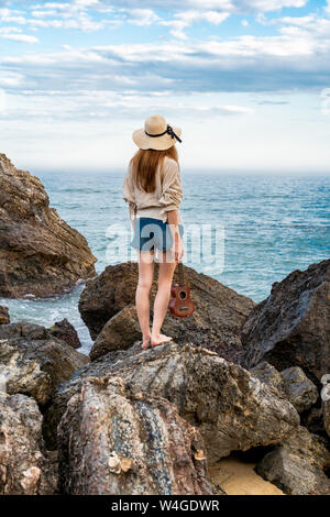 Vue arrière de jeune femme avec ukulele debout sur la roche face à la mer Banque D'Images
