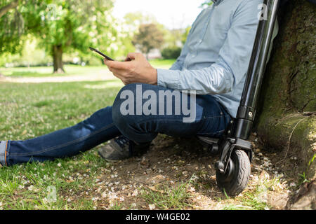 Homme assis sur l'E-scooter dans un parc leaning against tree trunk lors de l'utilisation de smartphone, vue partielle Banque D'Images